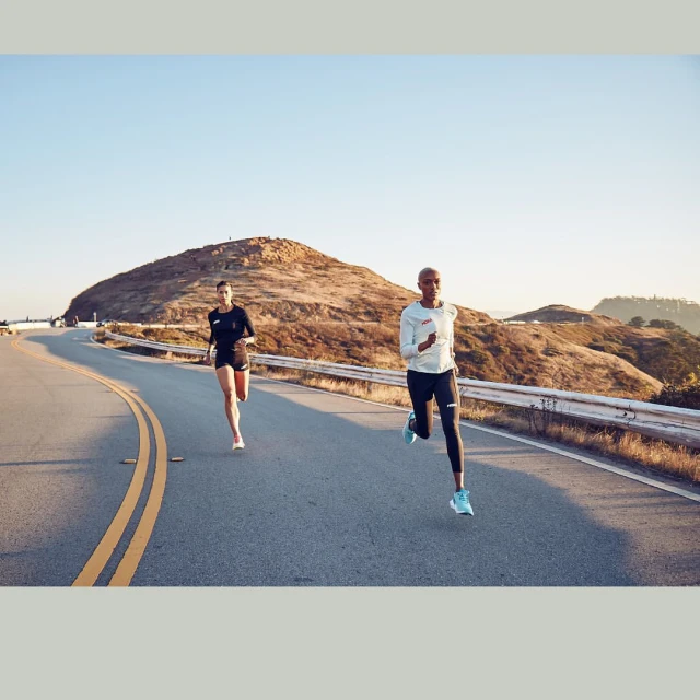 Two women run side-by-side on an open road with hills and a clear sky in the background, captured in broad daylight.