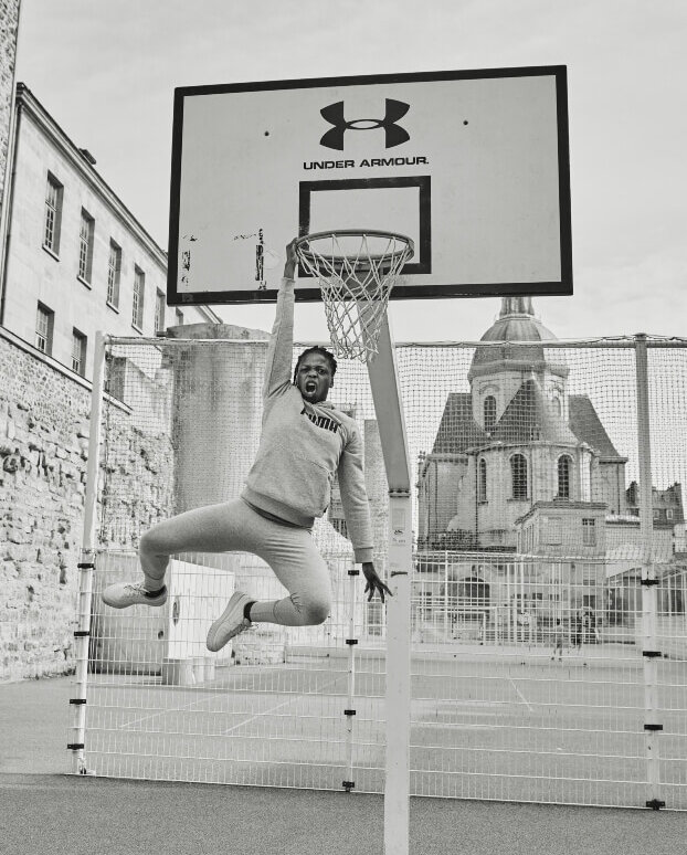Monochrome shot of an adult male athlete mid-air, grabbing a basketball hoop outdoors with architectural background.