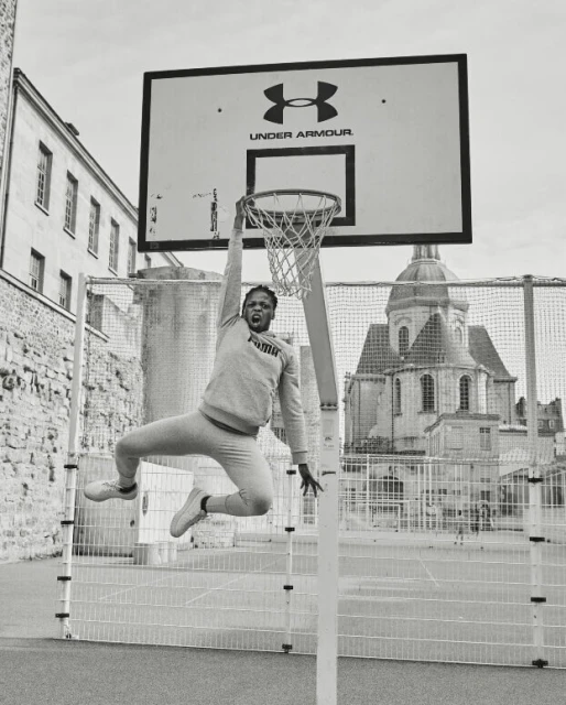 Monochrome shot of an adult male athlete mid-air, grabbing a basketball hoop outdoors with architectural background.