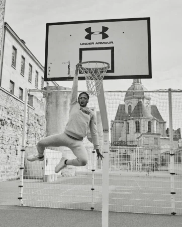 Monochrome shot of an adult male athlete mid-air, grabbing a basketball hoop outdoors with architectural background.