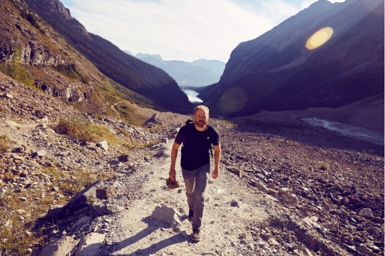 Man hiking on a rocky trail in a mountain landscape with a lake visible in the distance, wearing casual clothes and carrying a hat.