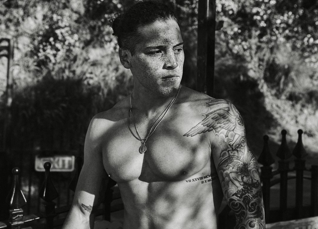 Monochrome portrait of a tattooed man with freckles and dark hair tied back, wearing a pendant necklace.