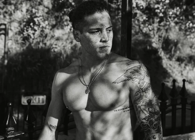 Monochrome portrait of a tattooed man with freckles and dark hair tied back, wearing a pendant necklace.