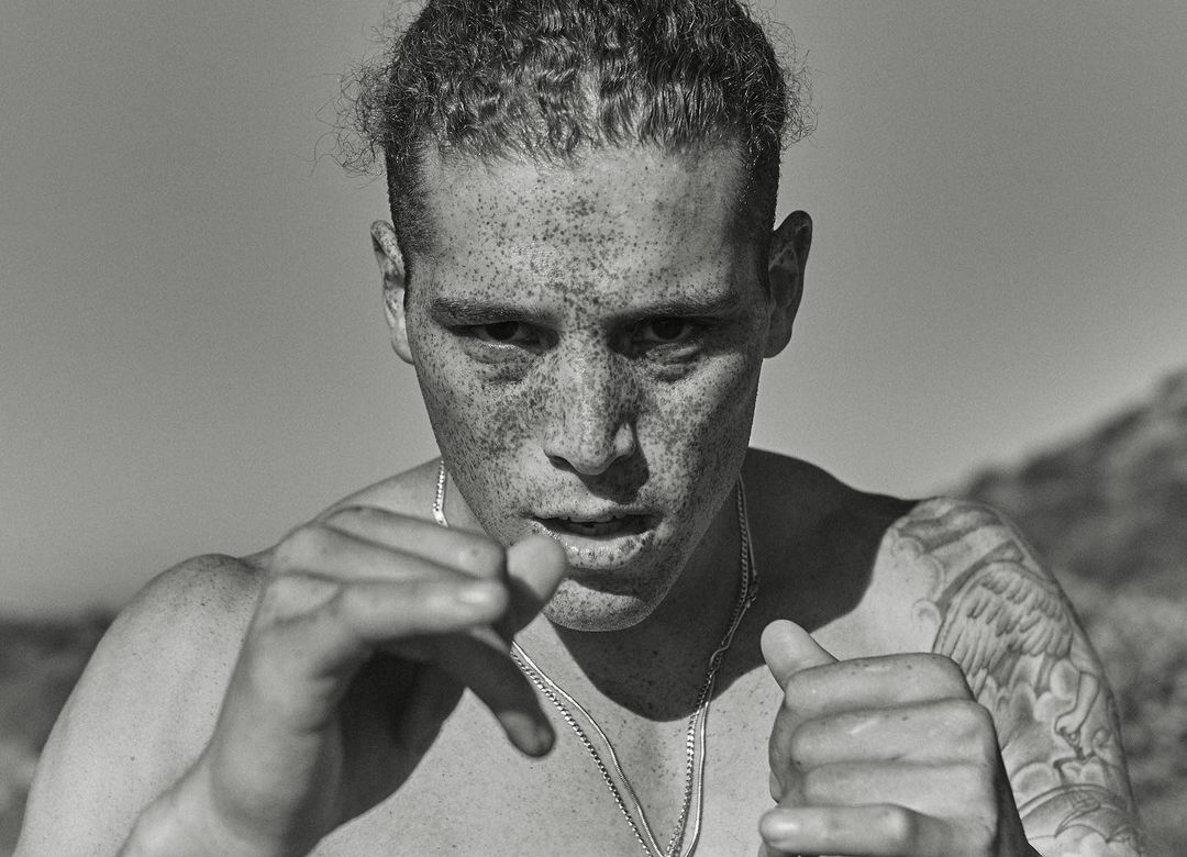 Monochrome portrait of a shirtless, freckled adult male in an outdoor setting, with a blurred background and a silver chain necklace.