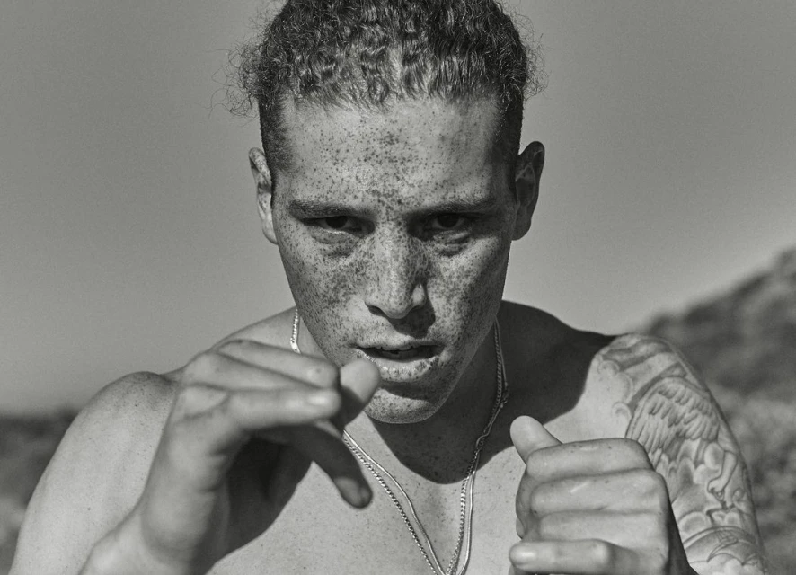 Monochrome portrait of a shirtless, freckled adult male in an outdoor setting, with a blurred background and a silver chain necklace.