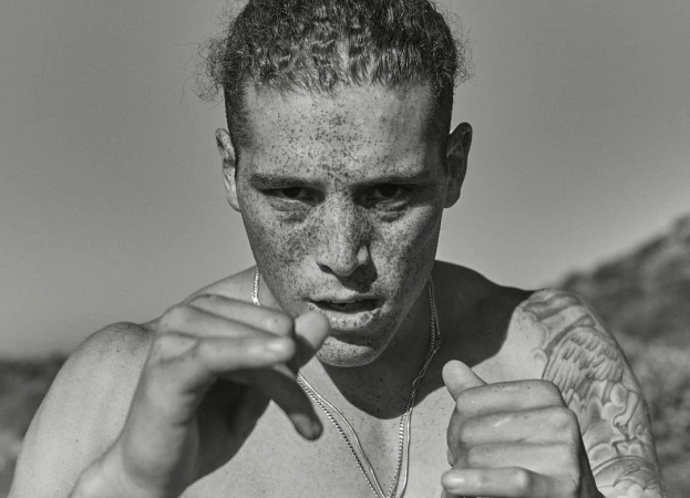 Monochrome portrait of a shirtless, freckled adult male in an outdoor setting, with a blurred background and a silver chain necklace.