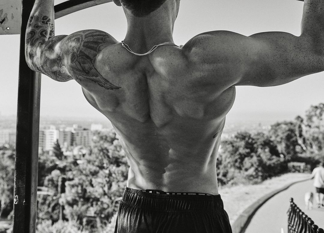 Muscular man with a tattoo doing pull-ups on a bar outdoors with a cityscape and greenery in the background, captured in black and white.