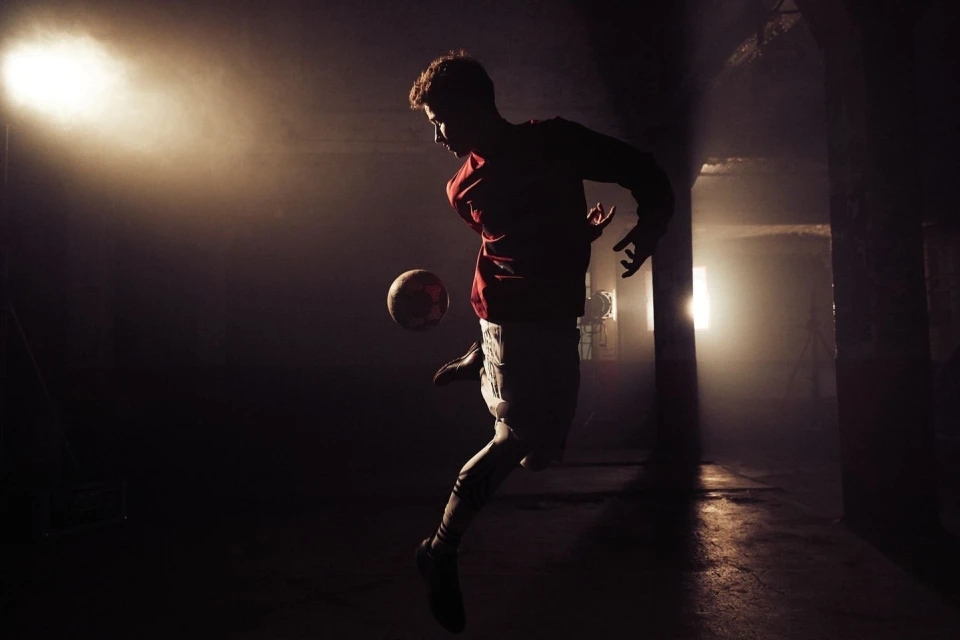 Athletic man in red shirt and white shorts juggling a soccer ball in a dark, foggy indoor training facility with bright backlighting.