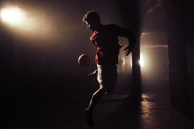 Athletic man in red shirt and white shorts juggling a soccer ball in a dark, foggy indoor training facility with bright backlighting.