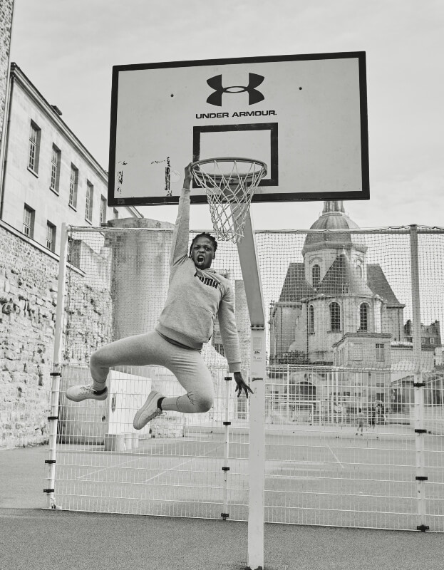 Monochrome shot of an adult male athlete in sportswear hanging from a basketball hoop on an outdoor court in an urban setting.