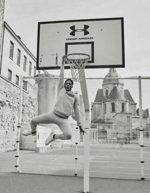 Monochrome shot of an adult male athlete in sportswear hanging from a basketball hoop on an outdoor court in an urban setting.
