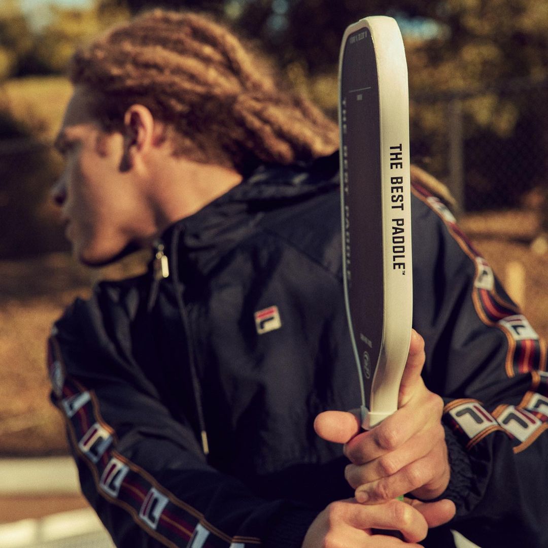 An adult male with dreadlocks is seen in a navy Fila jacket holding a pickleball paddle outdoors with natural light in the daytime.