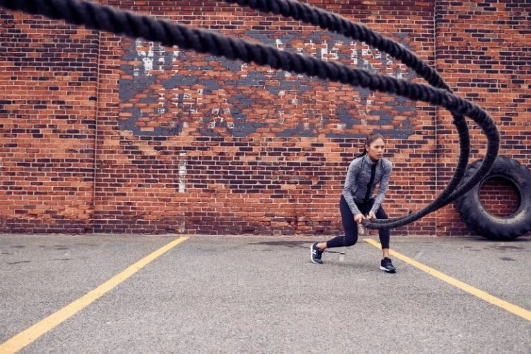 Athletic woman performs a battle ropes exercise outside, wearing sportswear in an urban setting with a brick wall and yellow lines.