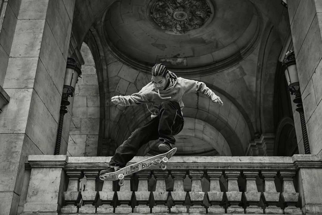 A skateboarder performs a trick off a ledge in an urban setting, with the backdrop of stone arches and vintage streetlights in monochrome.