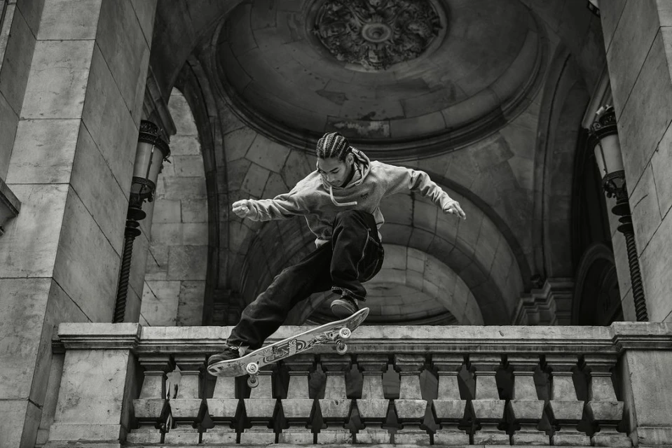 A skateboarder performs a trick off a ledge in an urban setting, with the backdrop of stone arches and vintage streetlights in monochrome.