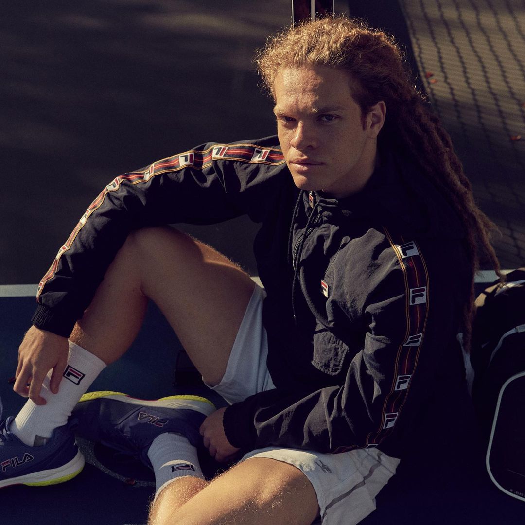 Athletic man with dreadlocks wears FILA jacket and shorts while sitting outdoors on a sunny day, showcasing sportswear fashion.