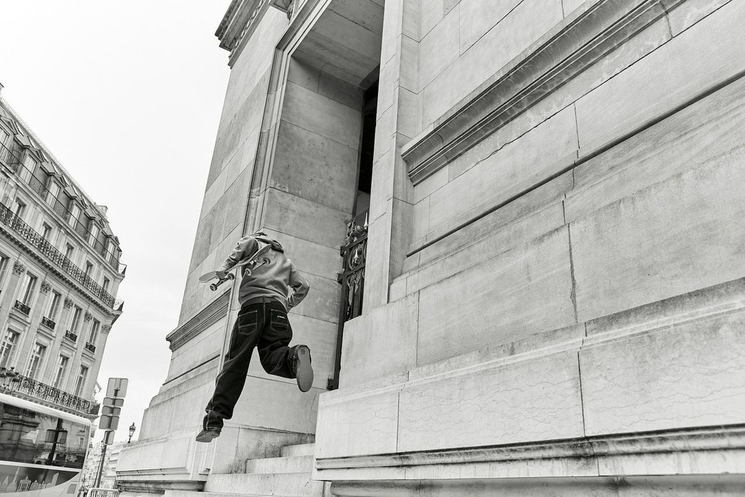 Person jumps stairs with a skateboard outside building in black and white, cityscape in the background, angled perspective, urban shot.