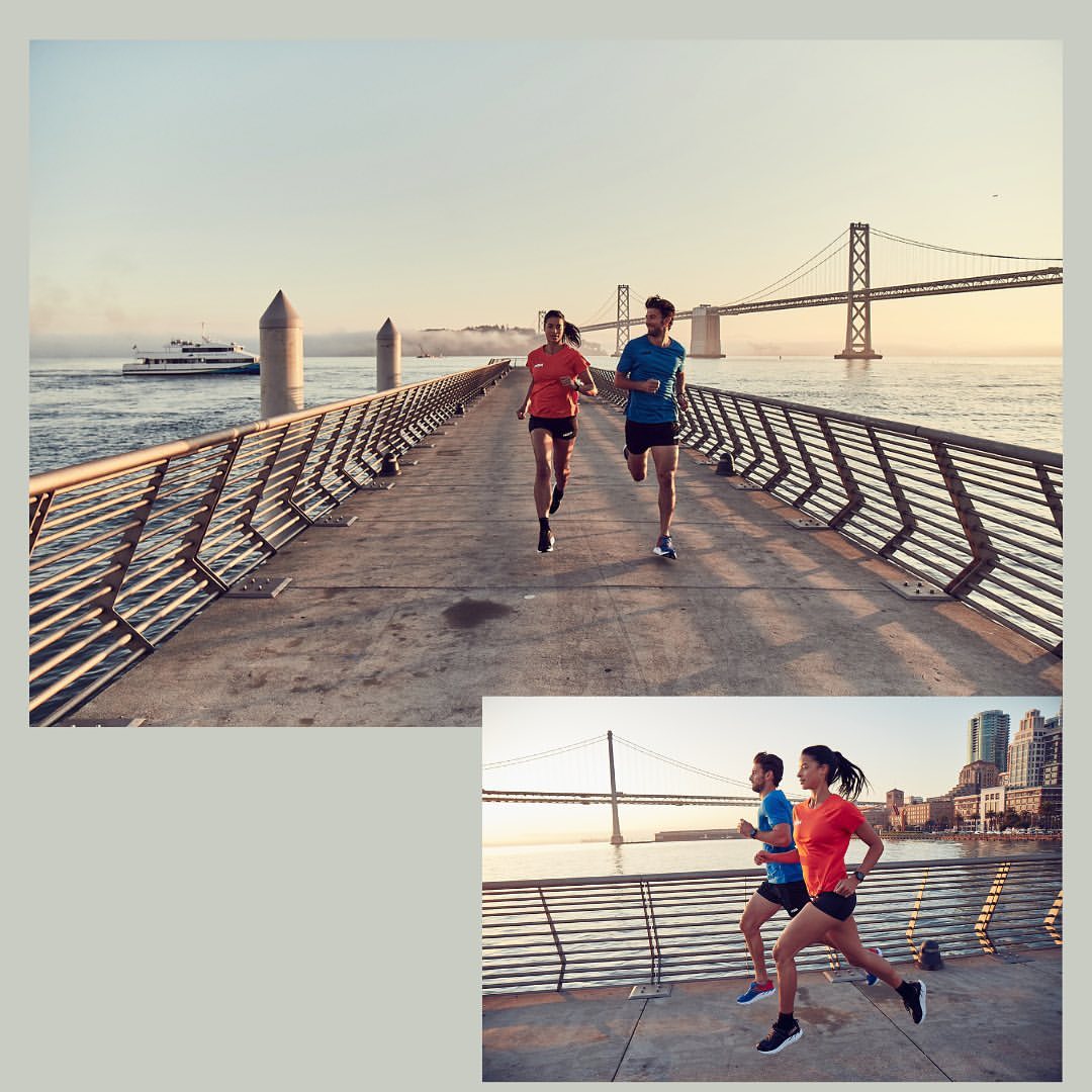 Two runners in activewear running along a pier with a large suspension bridge and boat visible in the background