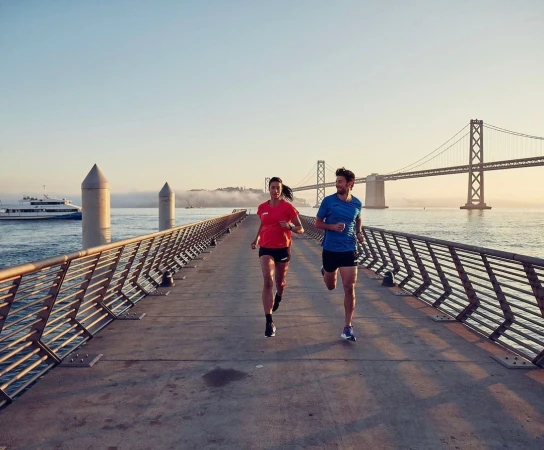 Man and woman running toward the camera along a concrete pier with a large suspension bridge visible in the background at sunset.