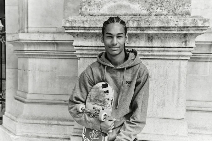 Monochrome portrait of a young man with braided hair wearing a hoodie and holding a skateboard, standing outdoors.