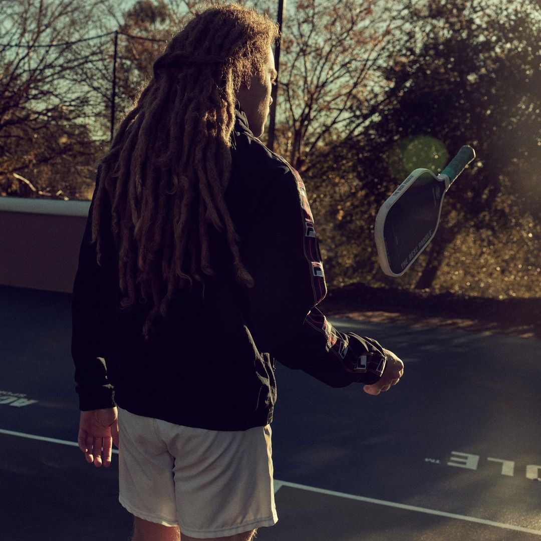 A man with dreadlocks outdoors wearing shorts and a jacket, tossing a pickleball paddle in the air, with trees and sunlight in the background.