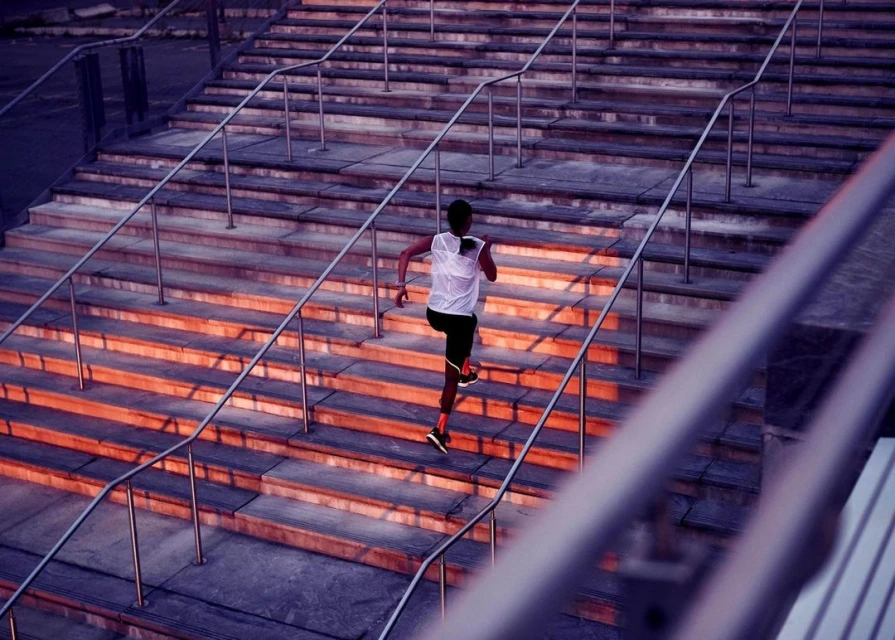 Athletic man in running attire ascends concrete stairs outdoors, illuminated by warm light creating dynamic shadows and texture.