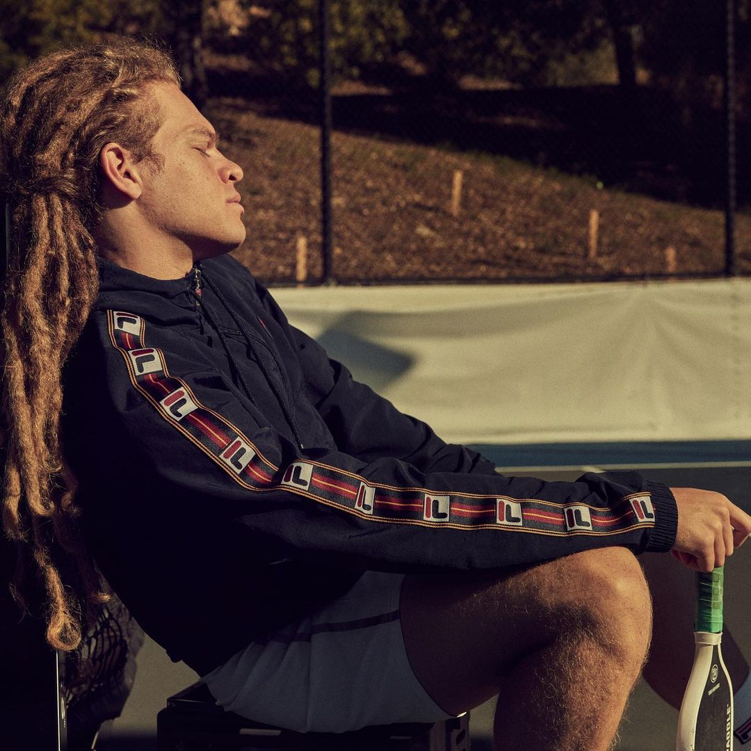 Adult man with dreadlocks sits outside, eyes closed, wearing Fila-branded jacket, shorts, and holding tennis racket, warm sunny lighting.