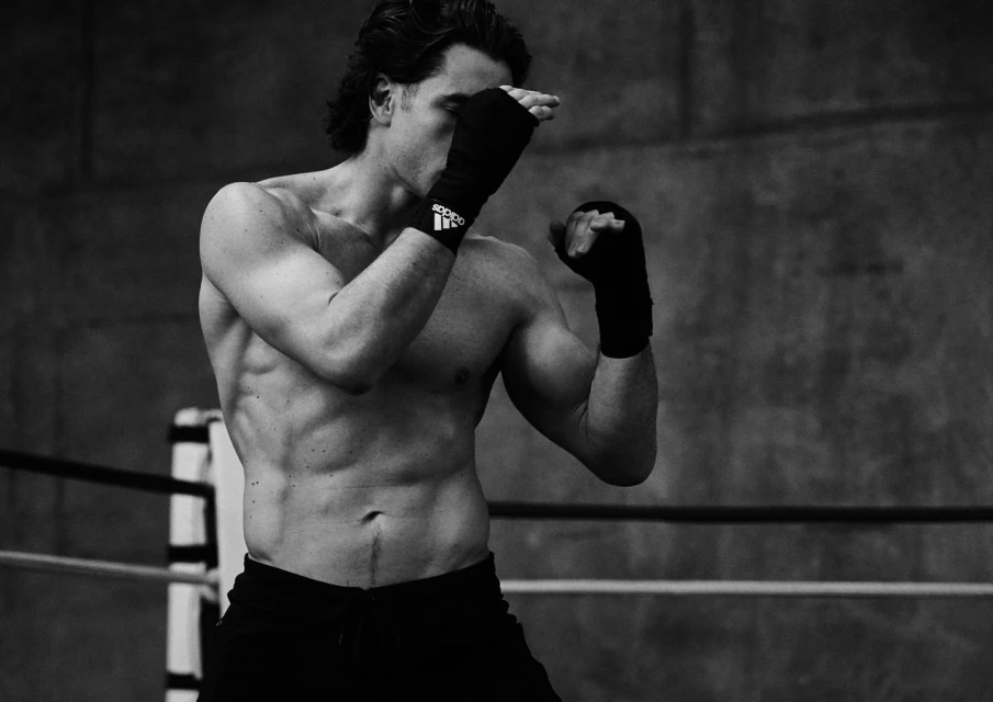 Black and white shot of a fit, shirtless adult male boxer with wrapped hands in fighting stance inside of a boxing ring.