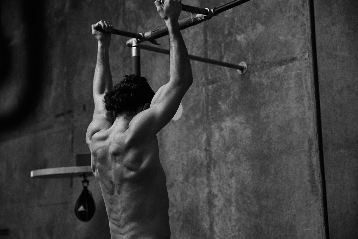 Shirtless adult male performs pull-up on metal bar against a concrete wall. Focus on musculature, black and white image, fitness theme.