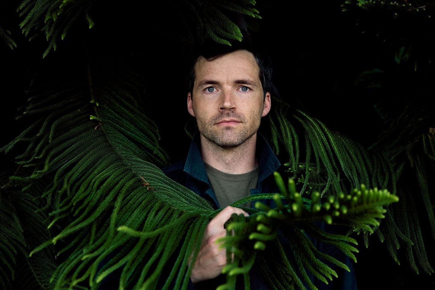 Close-up portrait of an adult man surrounded by lush green foliage in a dark, natural outdoor environment with soft, diffused lighting.