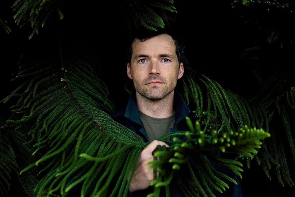 Close-up portrait of an adult man surrounded by lush green foliage in a dark, natural outdoor environment with soft, diffused lighting.
