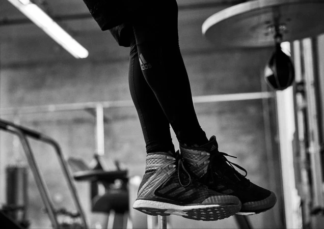 Monochrome shot of a person wearing Adidas leggings and high-top sneakers in a fitness gym, with a punching bag and exercise equipment visible.
