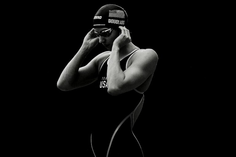 Portrait of female swimmer in swimsuit and cap adjusting goggles with a dark background in black and white, highlighting athletic physique.