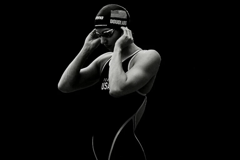 Portrait of female swimmer in swimsuit and cap adjusting goggles with a dark background in black and white, highlighting athletic physique.