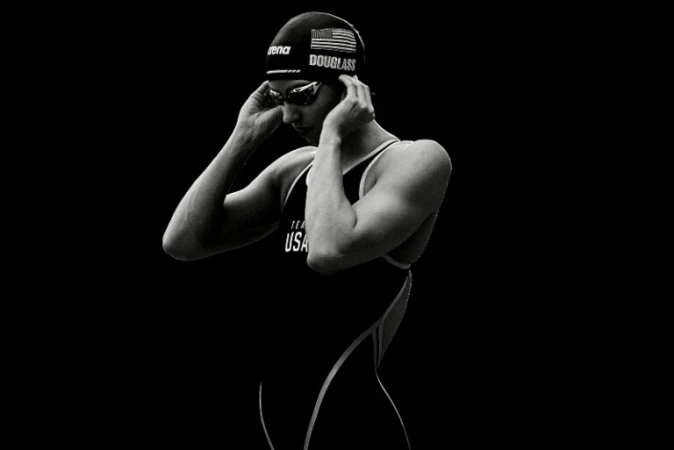 Portrait of female swimmer in swimsuit and cap adjusting goggles with a dark background in black and white, highlighting athletic physique.