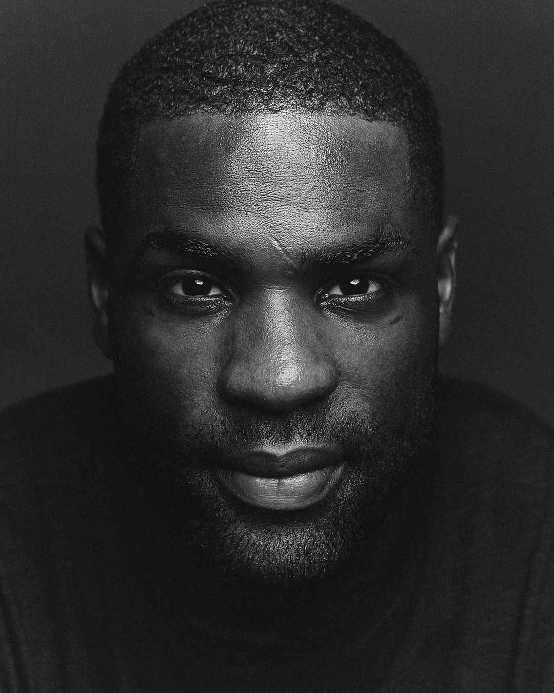 Black and white studio portrait of a serious adult male with short dark hair and a beard, looking directly at the camera.