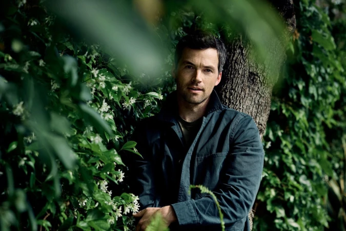 Man in a dark jacket posing outdoors among dense green foliage and flowering vines