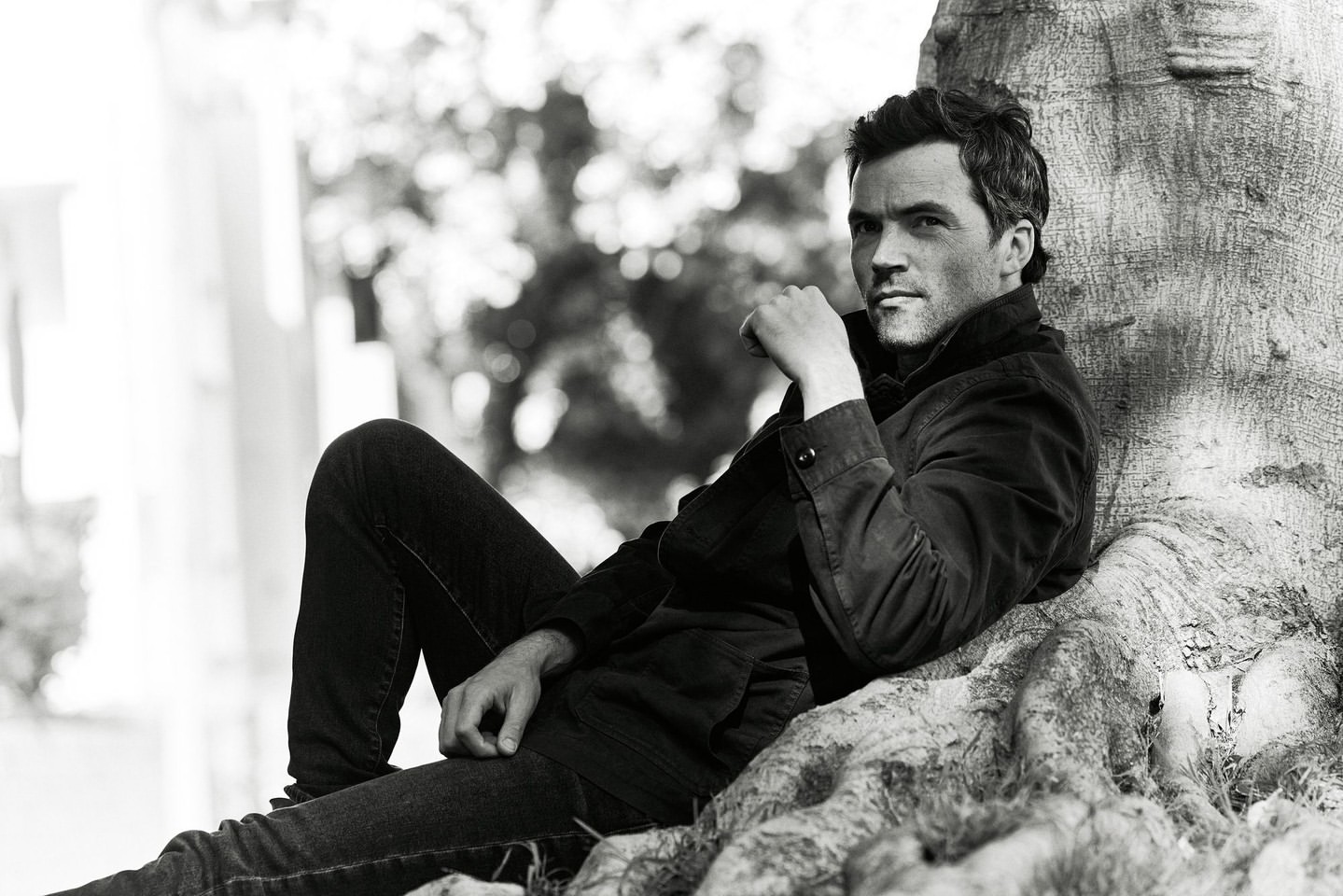 Man sitting against a tree in a black and white portrait. He wears a dark jacket and jeans, gazing thoughtfully towards the camera.