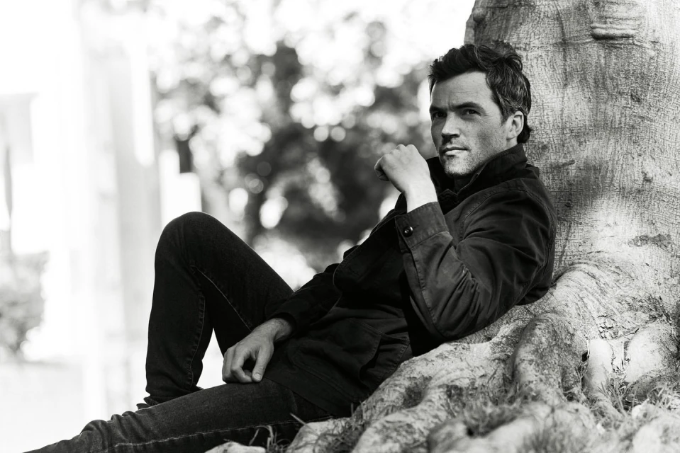 Man sitting against a tree in a black and white portrait. He wears a dark jacket and jeans, gazing thoughtfully towards the camera.