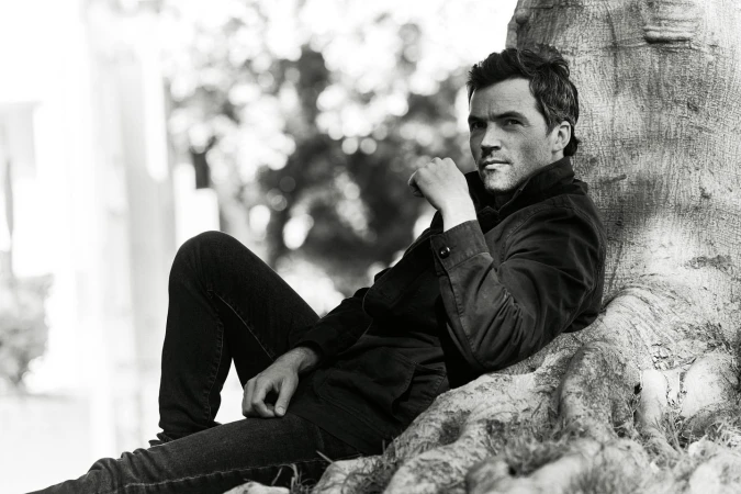 Man sitting against a tree in a black and white portrait. He wears a dark jacket and jeans, gazing thoughtfully towards the camera.