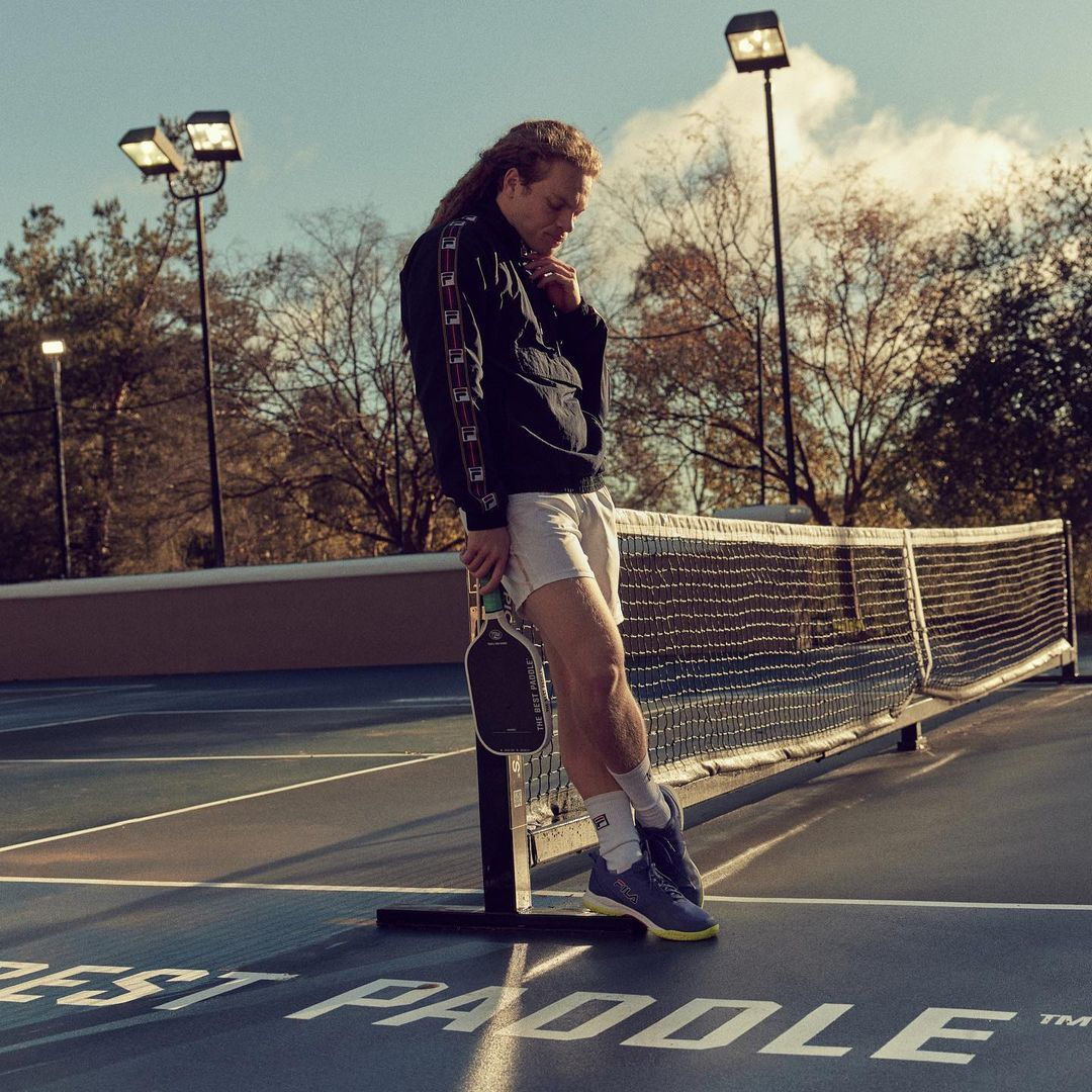 A man stands by a pickleball net, clad in a windbreaker and shorts, holding a paddle, photographed on an outdoor court on a sunny day.