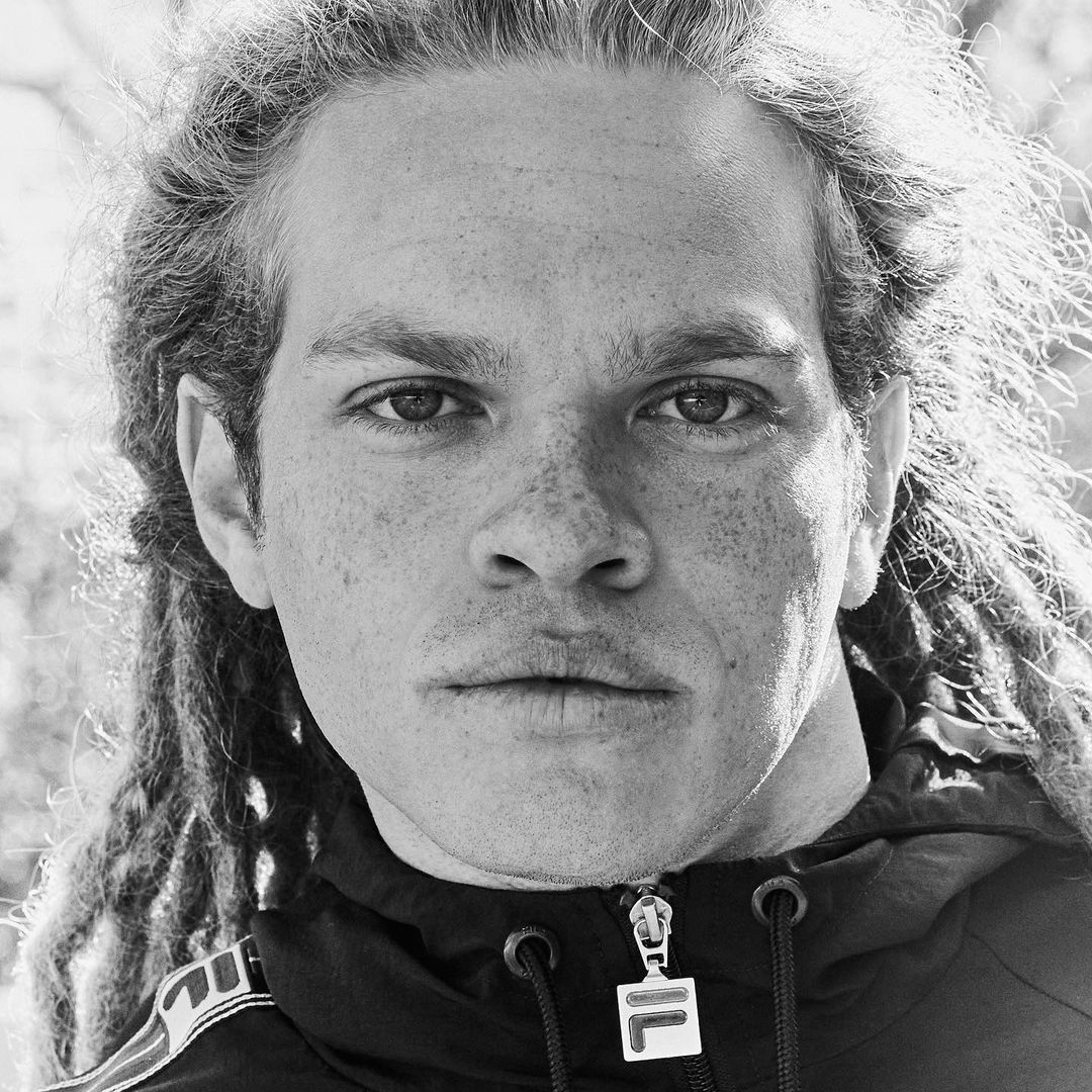Black and white portrait of a man with dreadlocks wearing a zip-up jacket, eyes looking directly at the camera, against an outdoor background.