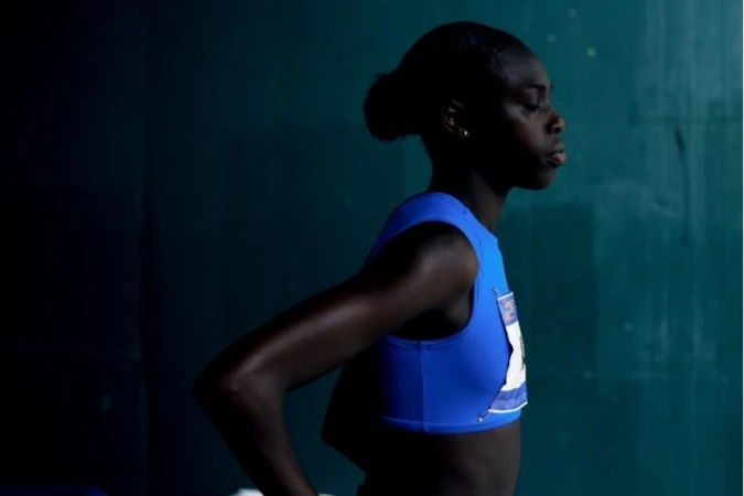 Black female athlete in blue sports top wearing a race bib standing against a dark green textured background.