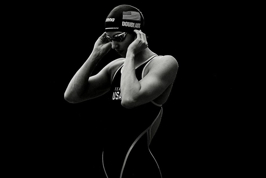 Black-and-white portrait of swimmer in swim cap and goggles, adjusting cap; wearing black swimsuit; dark, contrast-rich studio shot.