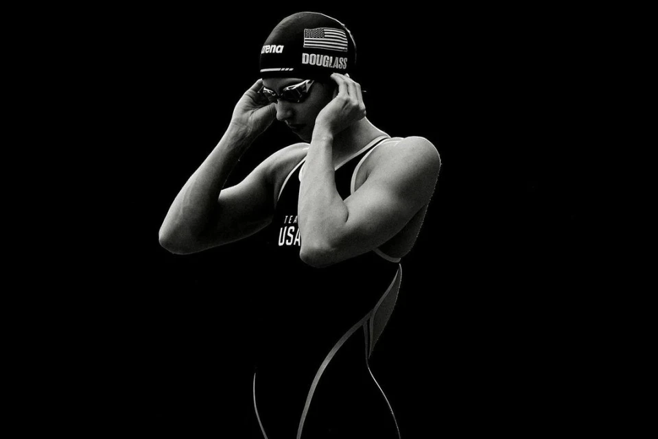 Black-and-white portrait of swimmer in swim cap and goggles, adjusting cap; wearing black swimsuit; dark, contrast-rich studio shot.