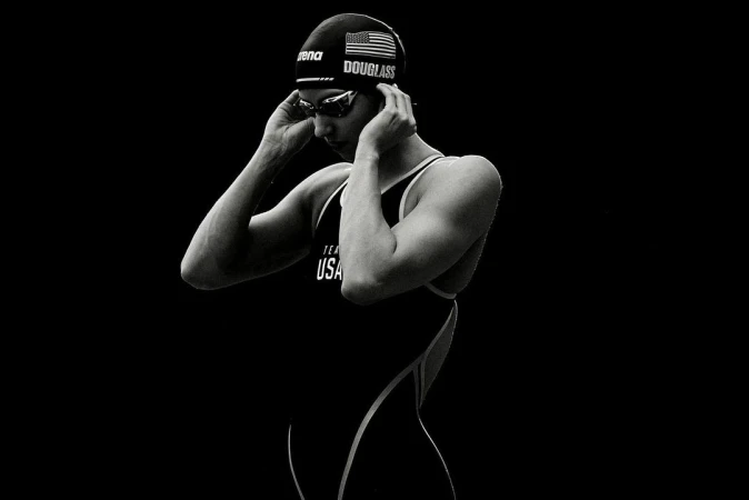 Black-and-white portrait of swimmer in swim cap and goggles, adjusting cap; wearing black swimsuit; dark, contrast-rich studio shot.