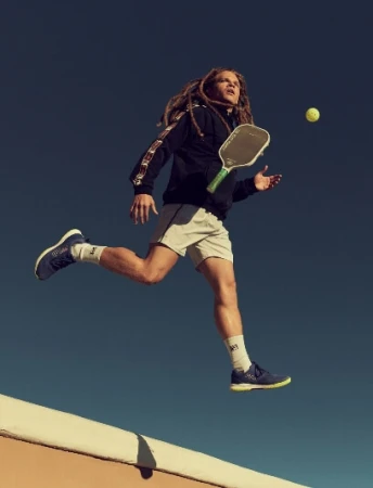 Man with dreadlocks mid-air playing pickleball wearing a black jacket and white shorts against a deep blue sky