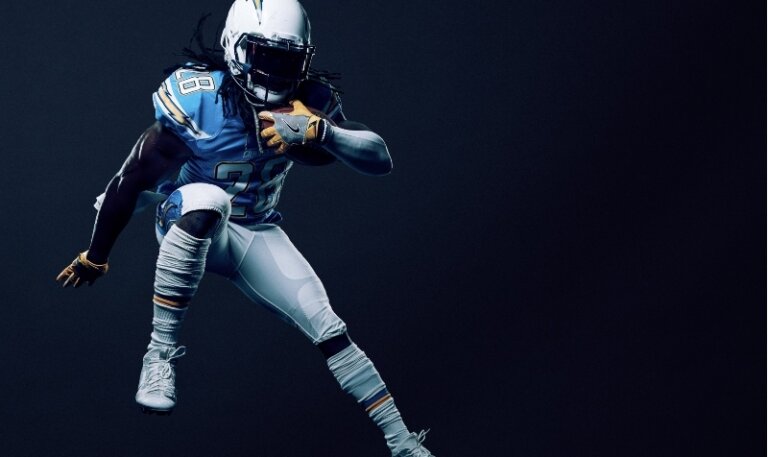 Action shot of a male American football player in blue and white uniform running with the ball against a dark blue background.