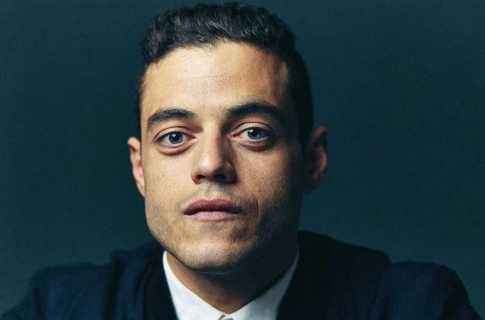 Close-up studio portrait of Rami Malek with a serious expression wearing a dark jacket in front of a dark blue background.