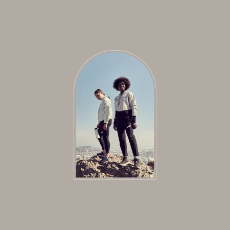 Two women stand on a rocky hilltop overlooking a city, wearing matching athletic outfits against a light blue sky framed by an arch.